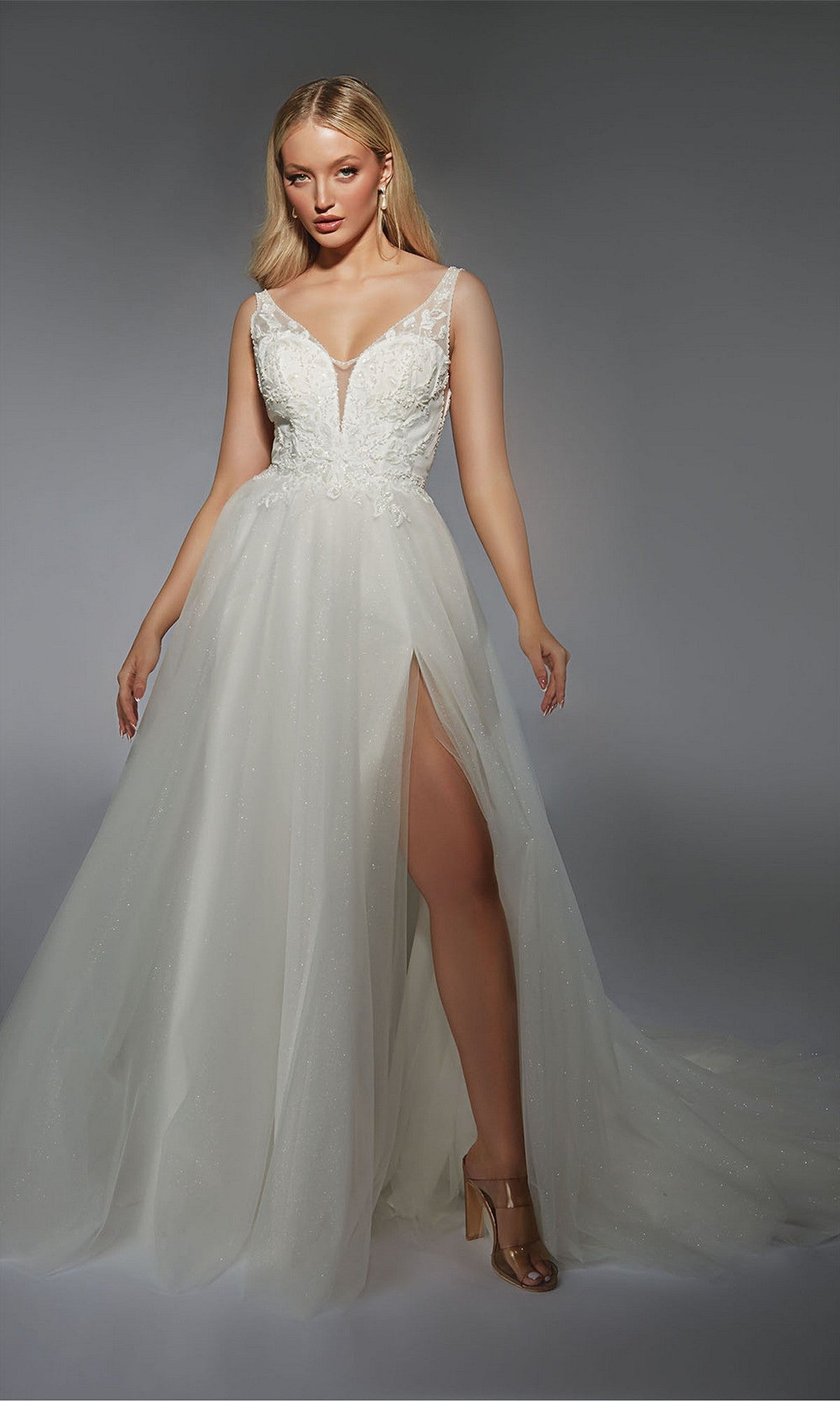 A woman models the Alyce 7125 Long White Dress, featuring a sleeveless design, deep V-neckline, floral bodice accents, and high leg slit—exuding elegant formal style against a plain gray background.